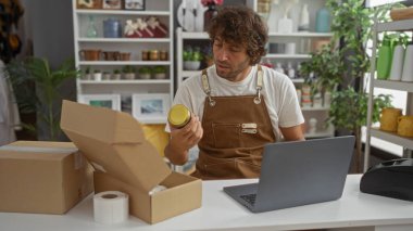Önlük giymiş genç bir adam dizüstü bilgisayarla evde oturuyor ve bir dekoru mağazasında eşyalar paketliyor.