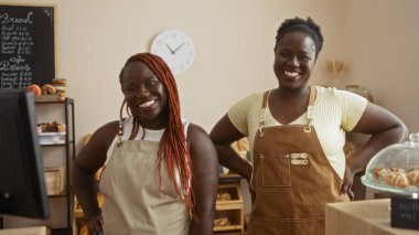 İki Afro-Amerikan kadın fırıncı sıcak bir pastanede gülümsüyor ellerinde hamur işleri ve ekmeklerle, içeride, arkalarında bir saatle..