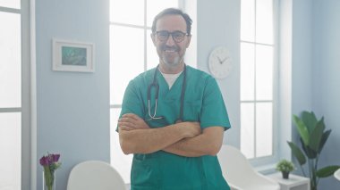 Yeşil önlüklü yakışıklı bir İspanyol erkek doktor, beyaz sandalyeli, çerçeveli sanat eserleri olan, parlak bir klinik lobisinde kollarını kavuşturarak duruyor..