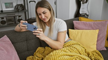 Kanepede uzanan genç bir kadın akıllı telefonunu karıştırıyor, rahat bir ortamda..