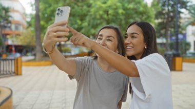 Şehir parkında akıllı bir telefonla selfie çekerken anne ve kızı gülümsüyor ve işaret ediyor..