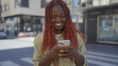 Neşeli genç bir Afrikalı kadın güneşli bir caddede akıllı telefon kullanıyor..