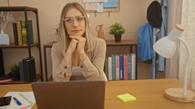 Genç, sarışın bir kadın, kitap, defter ve dizüstü bilgisayarla çevrili ev ofisinde düşünceli bir şekilde, modern bir çalışma alanı sunuyor..