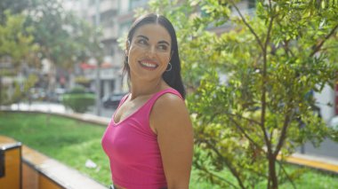 Pembe bluzlu bir kadın, güneşli bir günde şehir parkında yeşillik ve şehir binalarıyla gülümsüyor.
