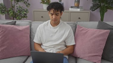 Genç İspanyol bir adam, renkli yastıklar ve iç mekan bitkileriyle çerçevelenmiş modern bir oturma odası ortamında dizüstü bilgisayarda çalışıyor..