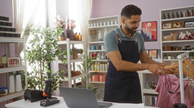 Önlüklü genç adam çeşitli dekorasyon malzemeleriyle şık bir mağazada kıyafet düzenliyor..