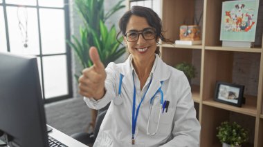 Gözlüklü, tıp önlüklü gülümseyen bir kadın klinikte başparmağını kaldırıyor..