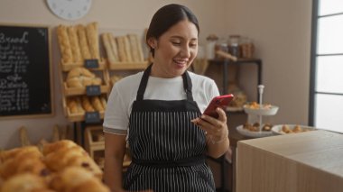 Fırında telefona bakarken gülümseyen bir kadın. Arkaplanda ekmek rafları ve hamur işleriyle.