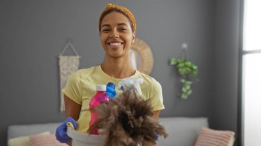 Genç bir kadın oturma odasını temizlerken elinde bir kova temizlik malzemesiyle gülümsüyor.