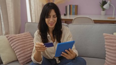 İnternetten alışveriş yaparken gülümseyen bir kadın tablet ve kredi kartıyla modern dekoratif ve doğal ışıklandırmalı rahat bir oturma odasında koltukta oturuyor..