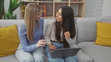 Kanepede dizüstü bilgisayar kullanırken, sıcak bir oturma odasında sohbet edip gülen iki kadın..