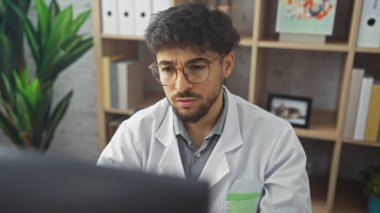 Gözlüklü konsantre Arap adam klinikte çalışıyor. Arka planda bilgisayar monitörü ve rafları var..