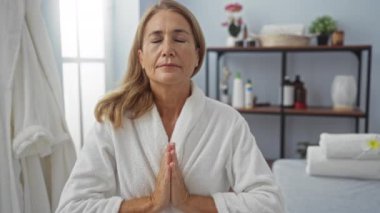 Beyaz cüppeli olgun bir kadın, sakin bir spa odasında meditasyon yapıyor, gözleri kapalı, sağlık ürünleri, çiçekler ve pencereden süzülen yumuşak ışıkla çevrili.