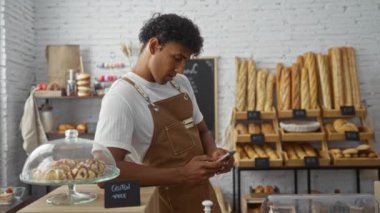Önlüklü genç adam fırında akıllı telefon kullanıyor. Etrafı ekmek ve pastalarla çevrili.