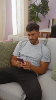 Oturma odasında telefon kullanan ve boynu ağrıyan genç bir adam modern bir ev ortamında ev hayatı ve genel sağlık sorunları sergiliyor gündelik giysiler giyiyor ve bir kanepede oturuyor.