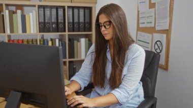 Ofisteki bilgisayarın başında çalışan genç bir kadın, profesyonel bir iş yerini, görevlerle meşgul esmer bir kadınla gösteriyor ve duygularını sergiliyor.