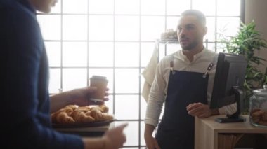 İki yetişkin adam bir pastanenin içinde etkileşime girer, biri tezgahın arkasında asistan olarak çalışır, diğeri de kahve fincanı tutarak siparişi tartışır..