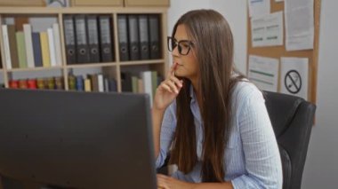 Ofis ortamında çalışan genç bir kadın, etrafı kitap ve kağıtlarla çevrili bir bilgisayar ekranına odaklanmış, profesyonel ve düzenli bir iş yerini ön plana çıkarıyor.