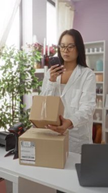 Akıllı telefon kullanan ve gözlük takan bir kadın dekore edilmiş bir ev içi mağazasında paketlerle uğraşıyor.