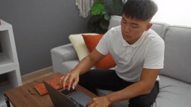 Stresli genç adam, modern bir oturma odasındaki bir kanepede otururken yüzünü kapatıyor, işinden bıkmış, açık bir dizüstü bilgisayar ve yakınlardaki masada dinlenen bardaklar gibi..