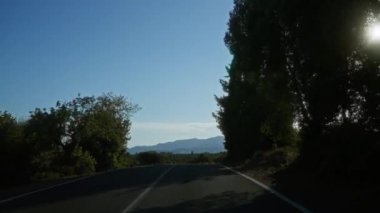 Boş, dolambaçlı bir köy yolu. Etrafı yemyeşil ağaçlar ve açık mavi gökyüzünün altındaki uzak dağlarla çevrili..