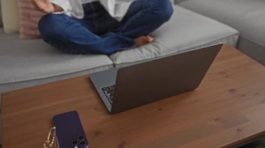 Kanepede meditasyon yapan bir kadın rahat oturma odasında dizüstü bilgisayar kullanıyor dekoratif minderlerle sakin ve rahat bir atmosfer sergiliyor.