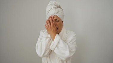 Havlu ve bornozlu bir kadın, yorgun bir şekilde esnerken yüzünün yarısını eliyle ve ağzıyla örtüyor, sabahın geç saatlerinde izole edilmiş beyaz arka planda yorgunluk çekiyor.