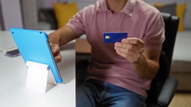 Evin içinde kredi kartı tutan ve oturma odasındaki bir masada tablet kullanan genç bir adam online alışveriş ya da finansal işlemleri gösteriyor.