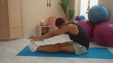 Spor salonunda egzersiz topları, mumlar ve arka planda bitkilerle esneyen genç bir adam esneklik ve sağlığa odaklanıyor.