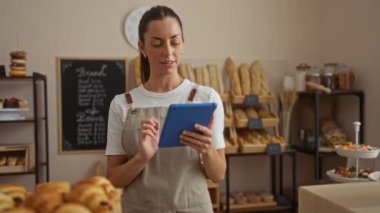 Fırında önlük giyen bir kadın tablet kullanıyor. Etrafı ekmek ve pastalarla çevrili.