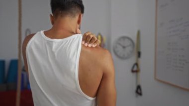 Spor salonunda omzunu tek eliyle tutan genç bir adam, arka planda egzersiz ekipmanıyla kapalı bir spor salonunda kas ağrısı ya da yaralanma belirtileri gösteriyor..
