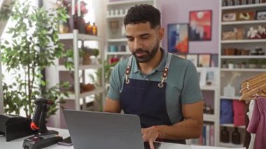 Önlüklü genç İspanyol adam dizüstü bilgisayar kullanıyor, baş parmağını kaldırmış, ev dekoratörü dükkanında ağzı açık gülümsüyor.