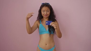 Bikini giyen ve pembe arka planda kredi kartına bakıp gülümseyen bir kadın.