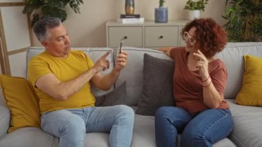 Orta yaşlı çift evde modern bir oturma odasında rahat bir koltukta otururken akıllı telefonlarıyla selfie çekiyorlar.
