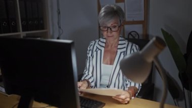 Ofiste gözlüklü, beyaz saçlı bir kadın bilgisayar monitörü ile gazete okuyor, gözle görülür şekilde stresli, masa lambasıyla aydınlatılıyor..