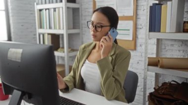 Ofis ortamında çalışan bir kadın, akıllı telefon ve bilgisayar kullanırken, etrafını kitaplık ve ilan panosuyla çevirmiş, odaklanma ve üretkenlik gösteren profesyonel görevlerle meşgul..