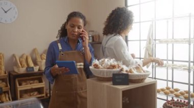 Bir fırında çalışan kadınlar, biri tablet ve telefon kullanıyor, diğeri ise raflardaki hamur işlerini düzenliyor, kapalı bir dükkânda pişmiş mallarla çevrili.