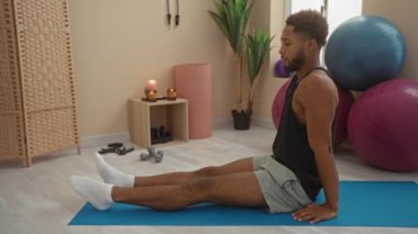 Spor salonunda spor salonunda spor yapan genç bir adam, etrafında spor aletleri olan, arka planda spor topları ve bitkilerle mavi yoga minderinde oturarak esneme hareketi yapıyor.