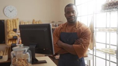Pastanedeki genç adam gülümsüyor, başparmağını kaldırıyor ve arka planda ekmek sergilenen hamur işleri ve kasanın yanında duruyor..
