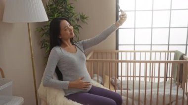 Arka planda lamba ve beşiğiyle rahat bir yatak odasında otururken selfie çeken hamile bir kadın.