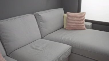 Genç adam gri kanepeli, pembe yastıklı, ev dekorasyonlu, günlük kıyafetler, doğal ışık ve şık dekorlu modern bir oturma odasında oturuyor..