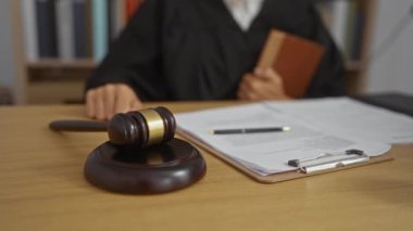 Hakim tokmağı mahkeme salonunda tutuyor, bornoz giyiyor, kitap tutuyor, masasında belgeler var.