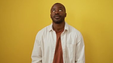 İzole edilmiş sarı arka planda duran, Afro-Amerikan tişörtlü, yukarı bakan, şaşırmış ve şaşırmış görünen bir adam.