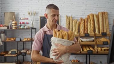 Kapalı bir pastanede elinde baget tutan genç adam çeşitli ekmek ve hamur işleriyle çevrili.