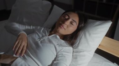 Kadın pijama giyerek yatakta geriniyor, rahat ve memnun görünüyor, yumuşak ışıklandırmalı rahat bir yatak odasında, huzurlu sabah rutinini ve sakin ev ortamını vurguluyor.