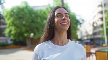 Arka planda ağaçlar ve binalar olan bir şehirde gündüz vakti şehir parkında gülümseyen genç bir kadın.