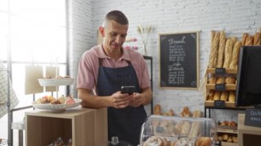 Fırın tezgahında akıllı telefon kullanan genç adam iyi aydınlatılmış bir dükkanda çeşitli hamur işleri ve ekmeklerle çevrili.
