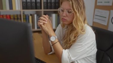 Sarışın kadın yorgun görünüyor ve bilgisayarı, kitapları ve klasörleri olan bir ofiste bardakları çıkarıyor..