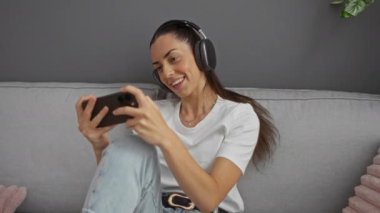 Akıllı telefonuyla oynayan, gülümseyen ve kulaklık takan açık renkli kot pantolon ve beyaz gömlekli bir kadın, rahatlamış, neşeli bir ifadeyle kapalı mekanda oyun oynuyor.