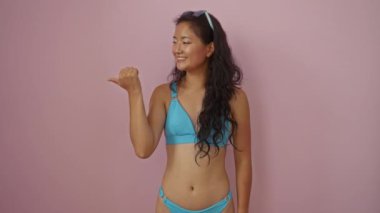 Bikinili genç bir kadın başparmağını kaldırmış, pembe arka planda ağzı açık gülümsüyor.